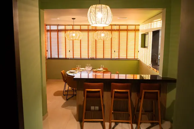 Sala de jantar com parede verde, bancada com banquetas, luminárias de fibra e persianas em tom madeira